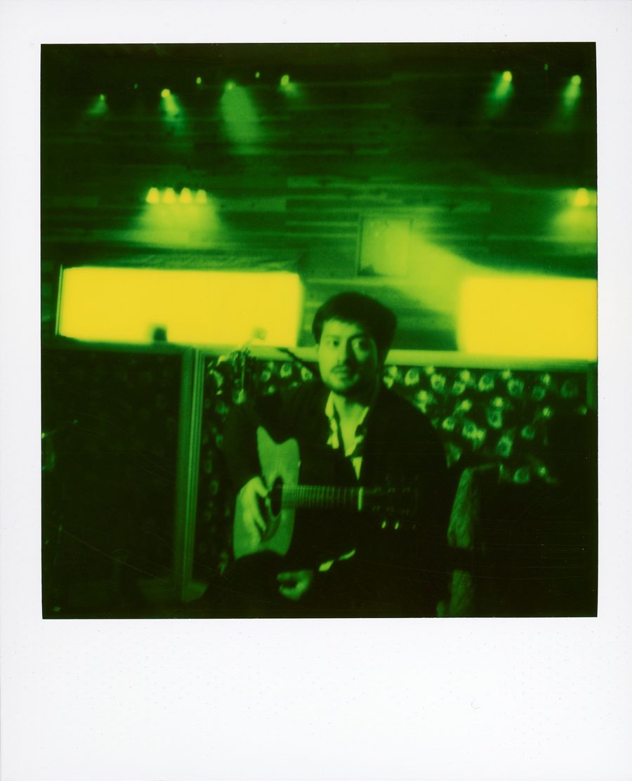 Polaroid verde de un músico con guitarra acústica en el estudio, con luces amarillas detrás
